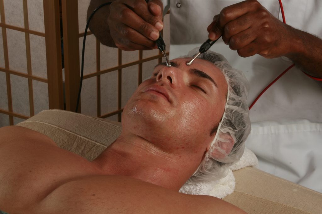 Man wearing a hair net receives a microcurrent facial treatment with probes applied to his face.